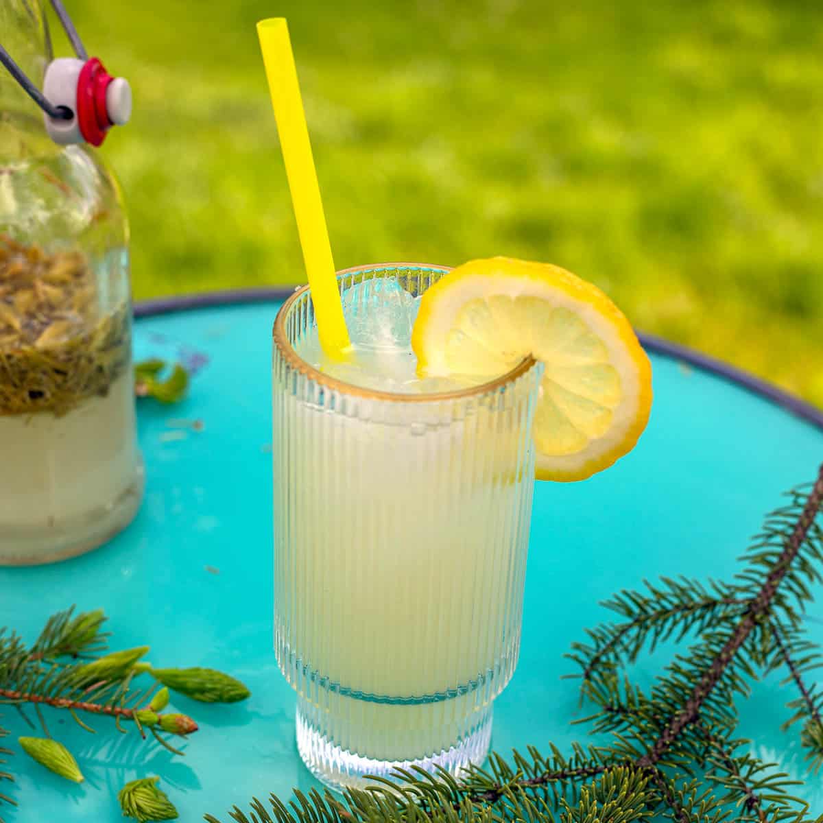 a glass of spruce tip soda with a yellow straw and a slice of lemon on a blue table.