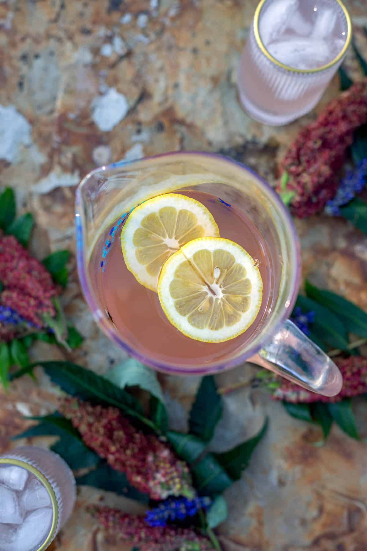 a pitcher of sumac lemonade with more sumac drupes and lavender scattered around it. 