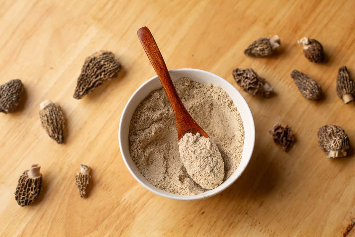 mushroom seasoning in a bowl with a wooden spoon and morel mushrooms scattered around it.
