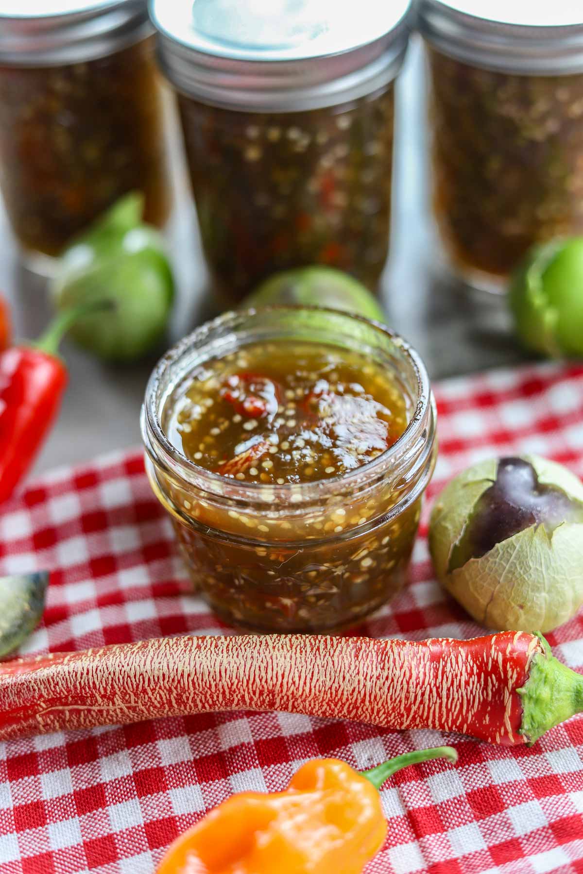spicy purple tomatillo jam in a jar with peppers and tomatillos around it and 3 filled jars behind it